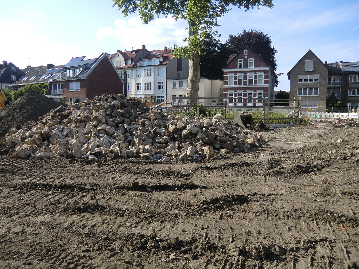 Bauschutt und Erde liegen vor einer begrünten Fläche mit modernen Häusern im Hintergrund